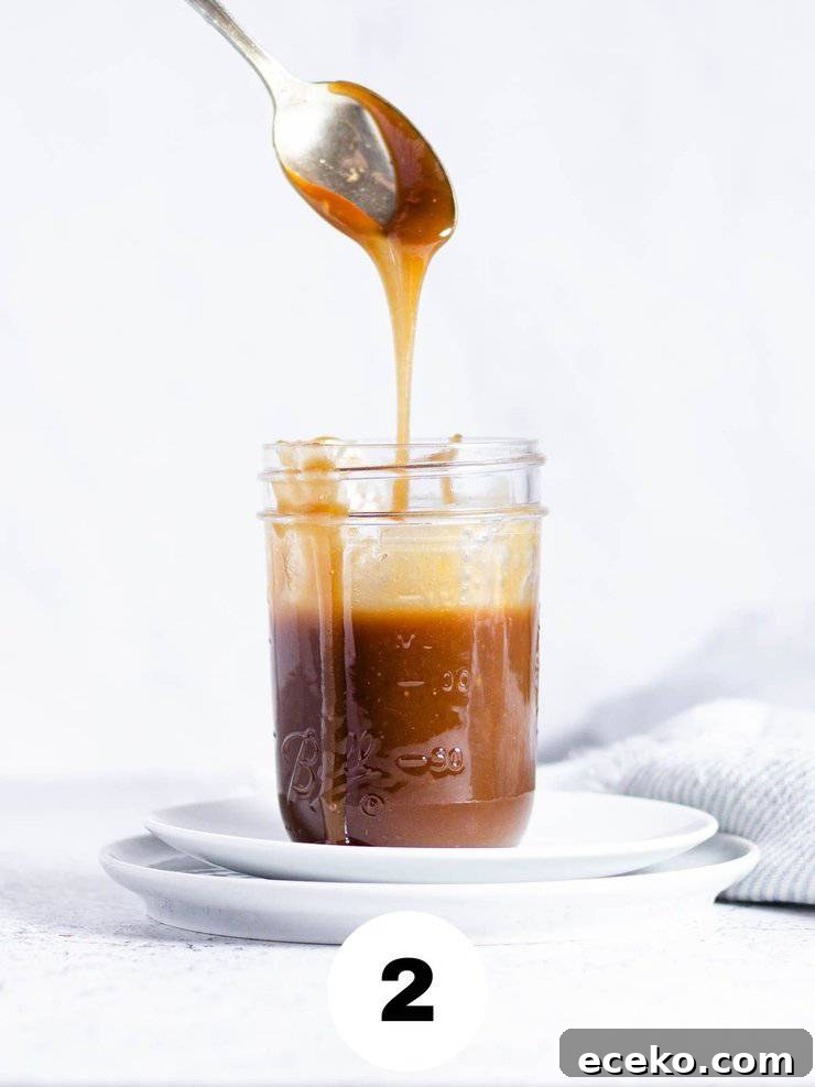 Side view of homemade caramel sauce dripping from a spoon into a clear mason jar.