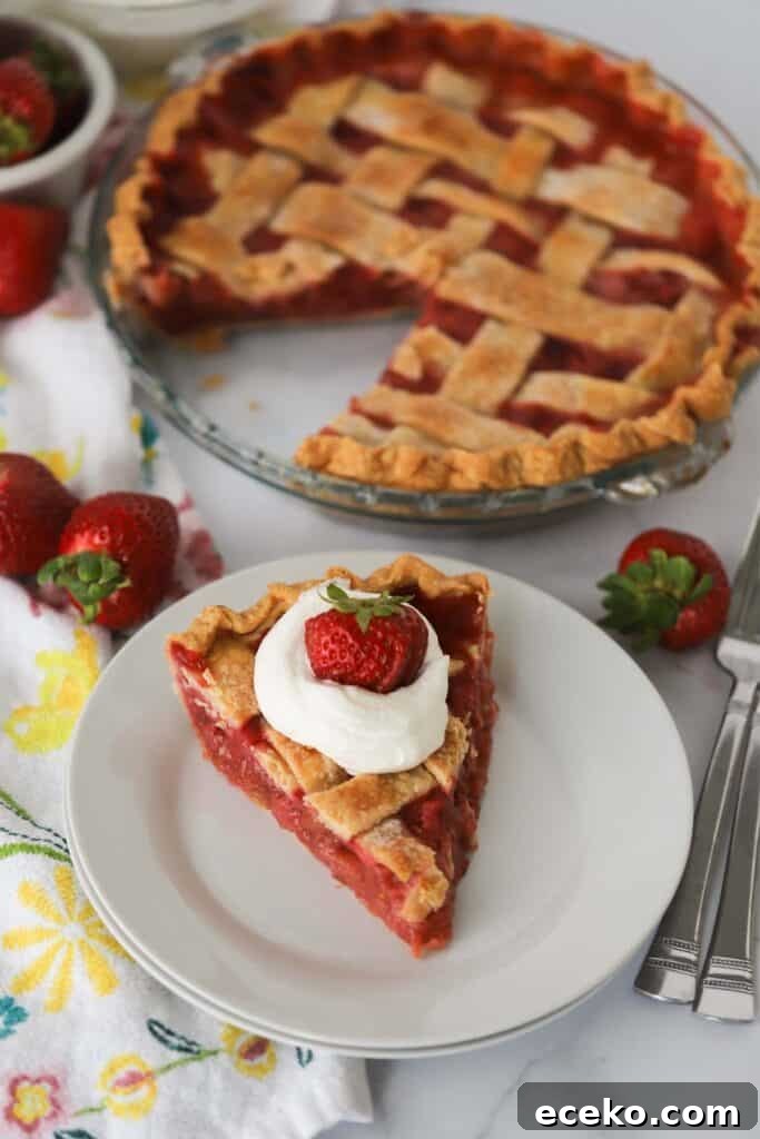 A slice of strawberry rhubarb pie on a white plate topped with whipped cream and a whole strawberry with a full pie in the background.