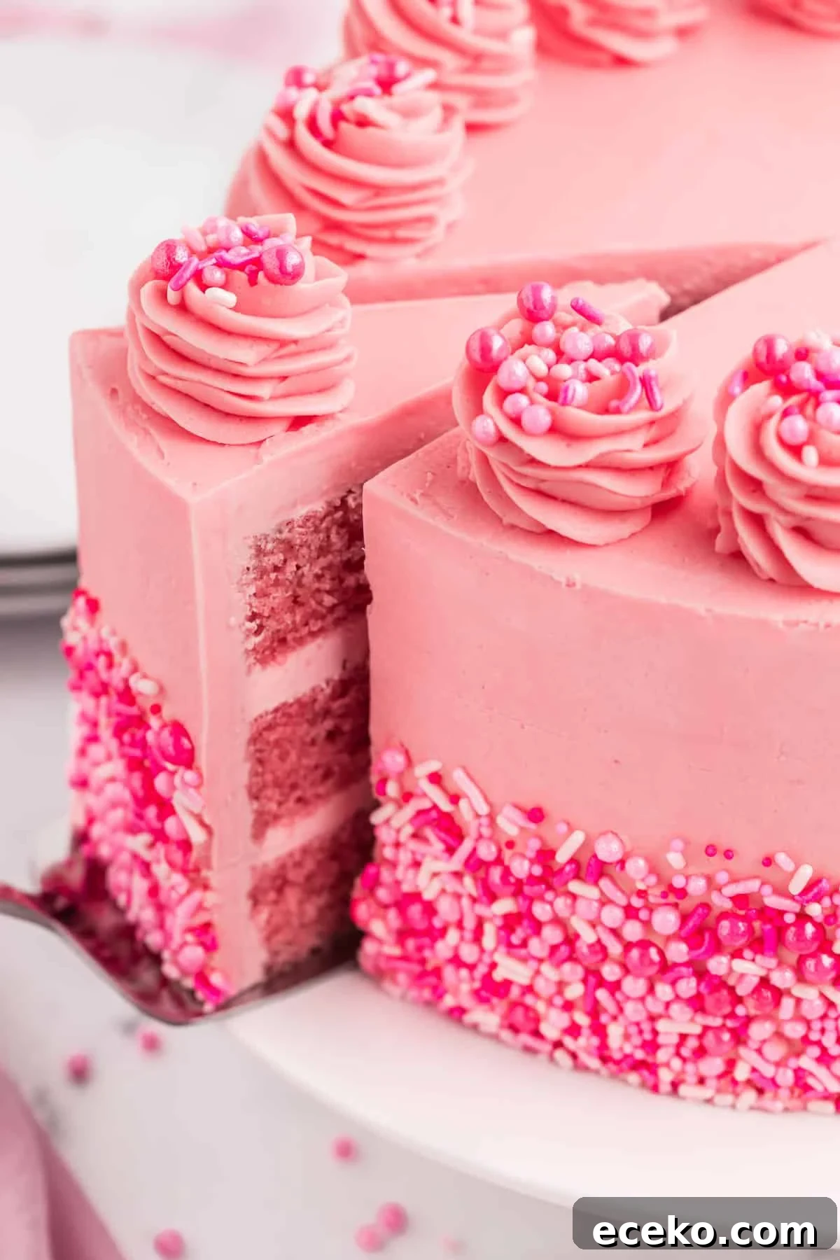 A pink cake with a slice being served out of it. The pink cake is decorated with sprinkles.