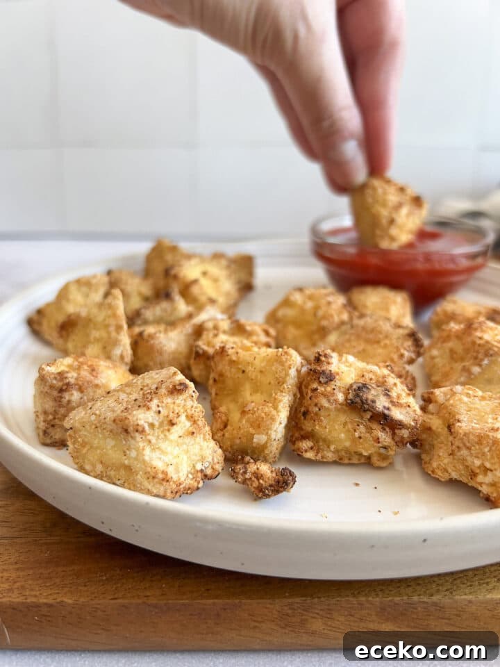 Crispy Air Fryer Tofu Nuggets — Quick Weeknight Recipe 10 Air fryer tofu nuggets on a white plate dipped, with a hand dipping a nugget in ketchup
