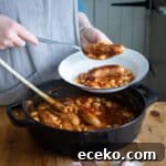 Cowboy Stew Recipes: Campfire, Stovetop, Oven & Slow Cooker Methods 17 womans hands dishing up cowboy sausage casserole from a black cast iron pan into a white enamel camping plate