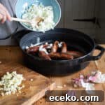 Cowboy Stew Recipes: Campfire, Stovetop, Oven & Slow Cooker Methods 19 woamns hands tipping chopped onions from a blue bowl into a black cast iron pan of cooked sausages
