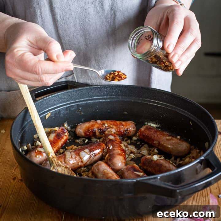 Cowboy Stew Recipes: Campfire, Stovetop, Oven & Slow Cooker Methods 6 Womans hands spooning chilli flakes from a small glass spice jar into a black cast iron pan of cowboy casserole