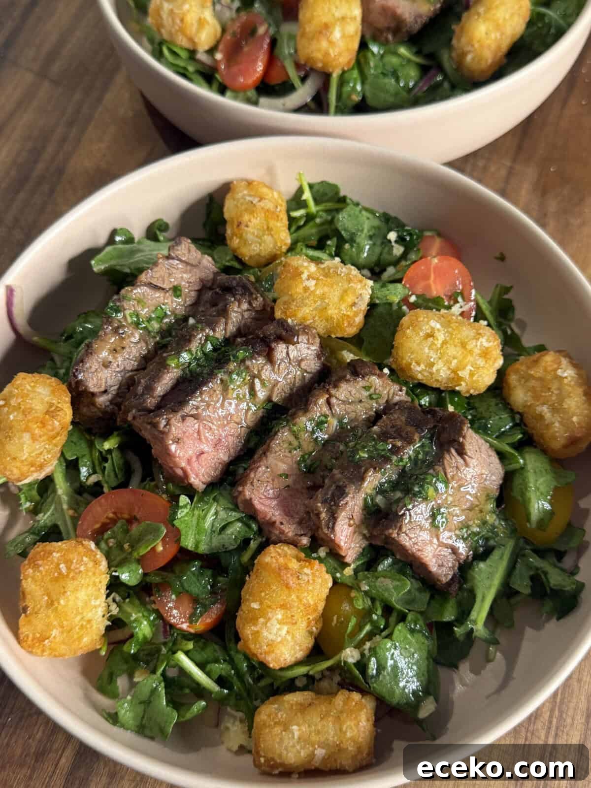 Steak and Potato Salad with Tangy Herb Dressing 2 A bowl filled with sliced steak on a bed of mixed greens, cherry tomatoes, red onions, and topped with golden tater tots, all set on a wooden table.