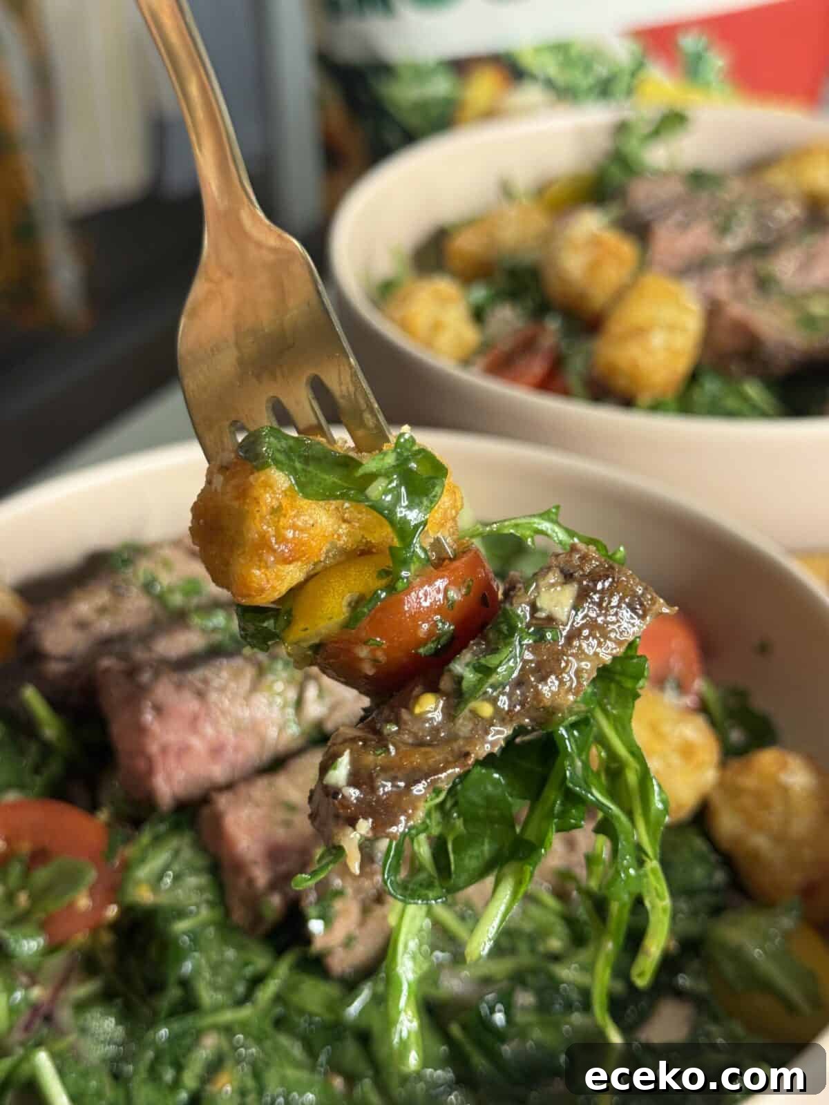 Steak and Potato Salad with Tangy Herb Dressing 3 A fork holds a bite of salad with arugula, cherry tomato, steak, and a crispy tater tot over a bowl filled with more steak, greens, and tater tots.