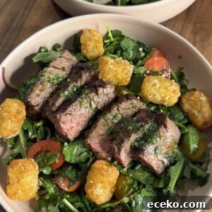 Steak and Potato Salad with Tangy Herb Dressing 4 A bowl filled with sliced steak on a bed of mixed greens, cherry tomatoes, red onions, and topped with golden tater tots, all set on a wooden table.