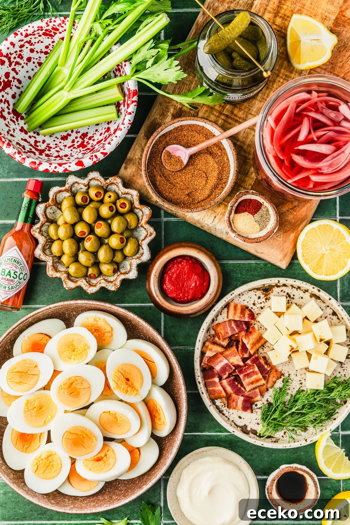 Brown bowls of hard-boiled eggs, bacon, cheese, dill, olives, tomato paste, Cajun seasoning, and spices next to a red and white bowl of celery, and jars of pickles and red onions on a green tiled counter.
