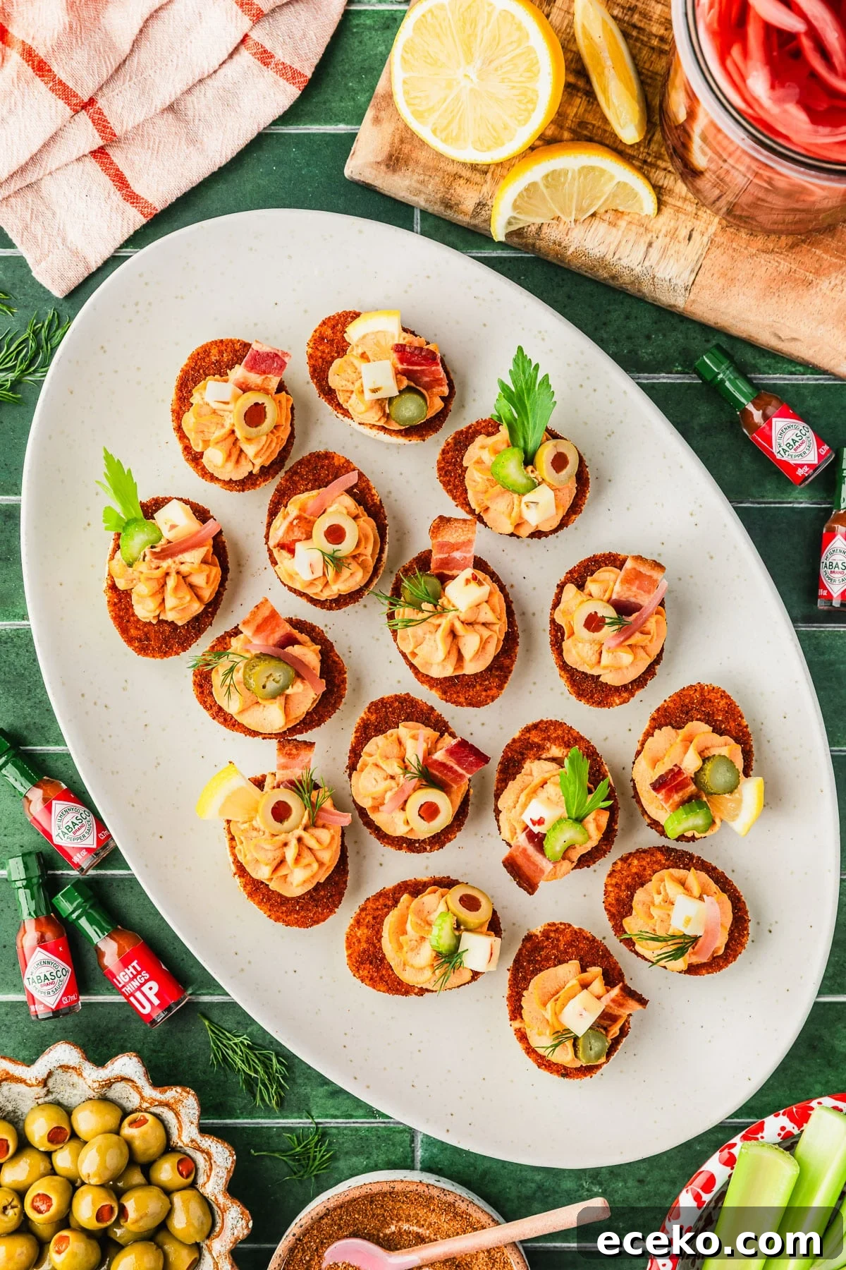 A white platter of Bloody Mary deviled eggs with bacon, celery, and other toppings on a green tiled counter. The platter is next to mini hot sauce bottles, a red and white bowl of celery, a jar of pickled onions, and brown bowls of olives and seasoning.