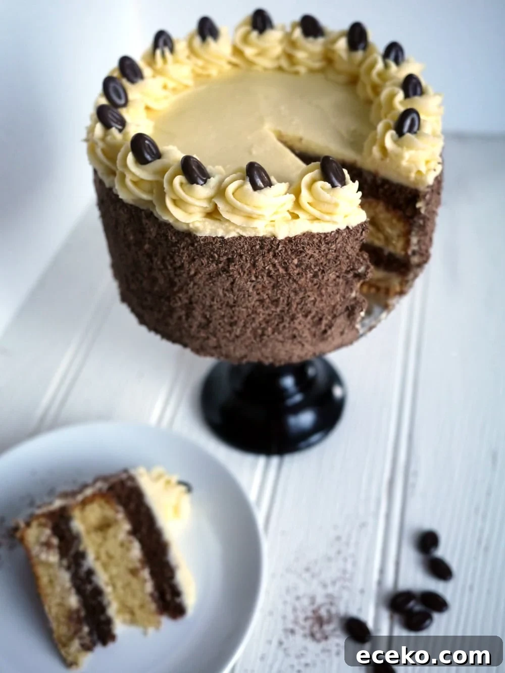 A layered Tiramisu Cake on a cake stand on a white surface with a portion missing, with a slice of cake on a plate at the side.