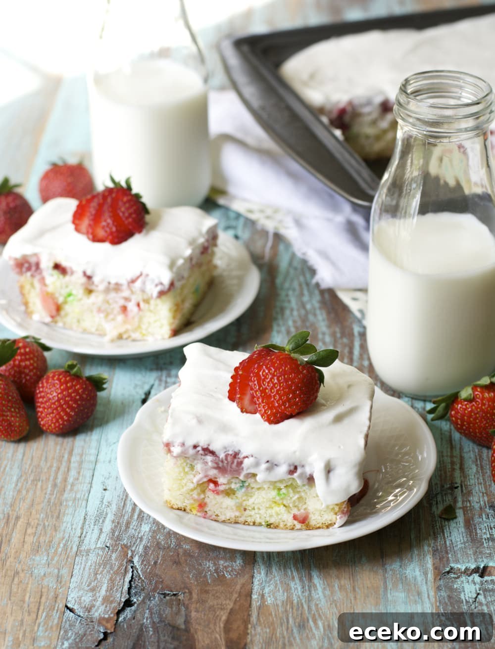 two slices of strawberry poke cake on white plates next to two glass jugs of milk. 