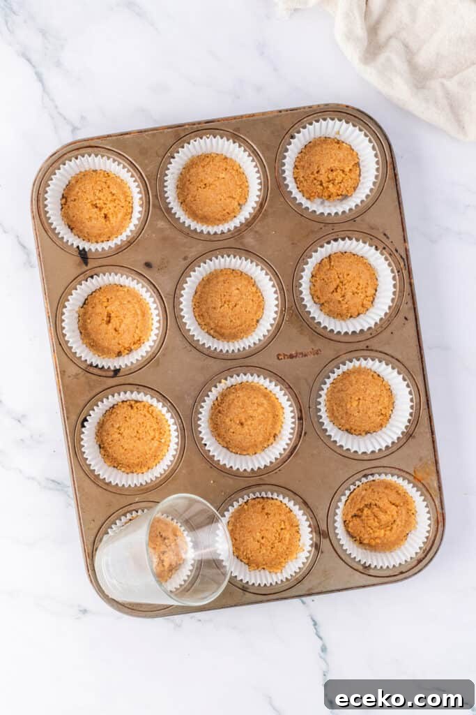 Mini No-Bake Chocolate Peanut Butter Cheesecakes 3 Muffin pan lined with white cupcake liners, each filled with pressed graham cracker crust. A small glass is shown pressing the crust into shape.