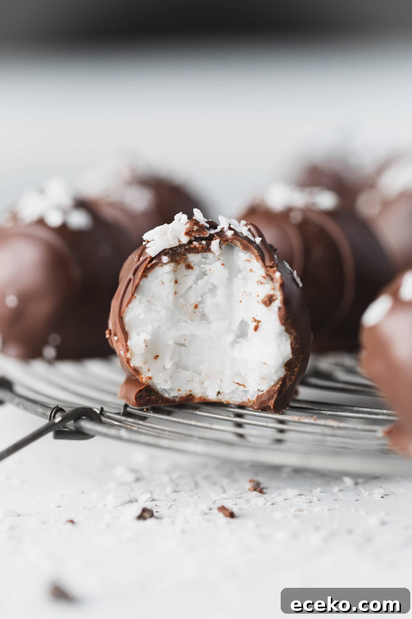A chocolate covered coconut ball that has been broken in half so you can see the creamy white coconut center. There are other coconut balls in the background.