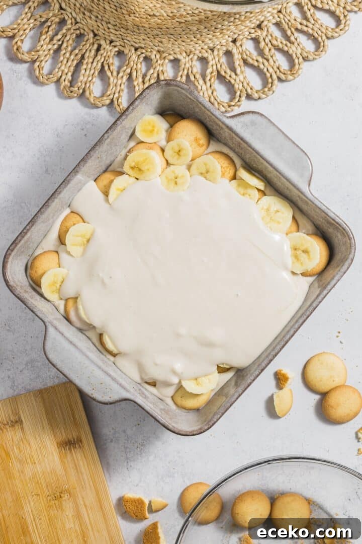 Banana pudding being assembled in a square baking dish with cookies and banana slices.