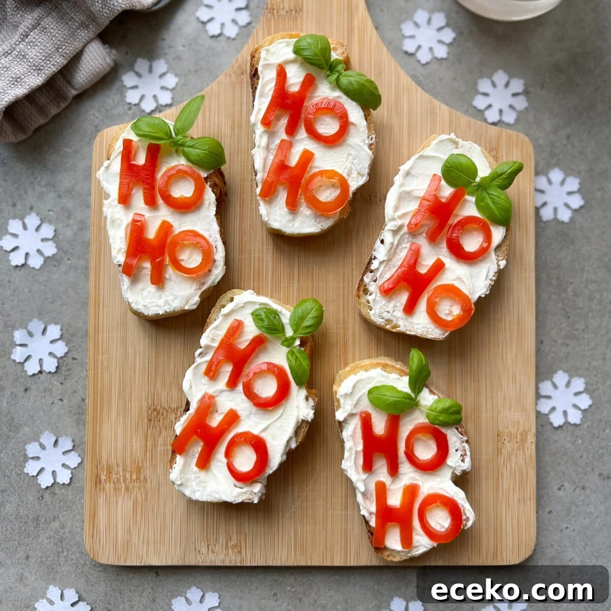 Festive Holiday Appetizers for Entertaining Guests 3 Five slices of Christmas Bruschetta with cream cheese, tomato letters spelling HO HO, and basil leaves are arranged on a wooden board with paper snowflakes on a grey surface.