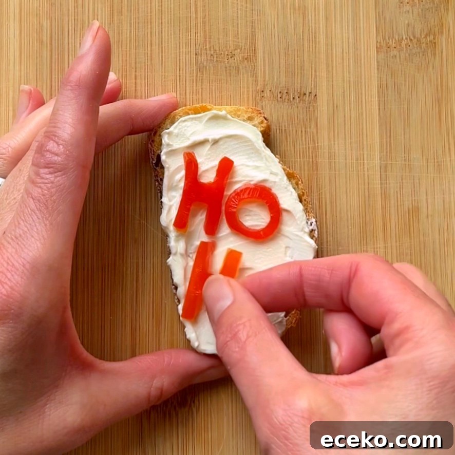Festive Holiday Appetizers for Entertaining Guests 6 Two hands decorate a piece of bread with cream cheese and tomato cutouts spelling HO on a wooden cutting board.