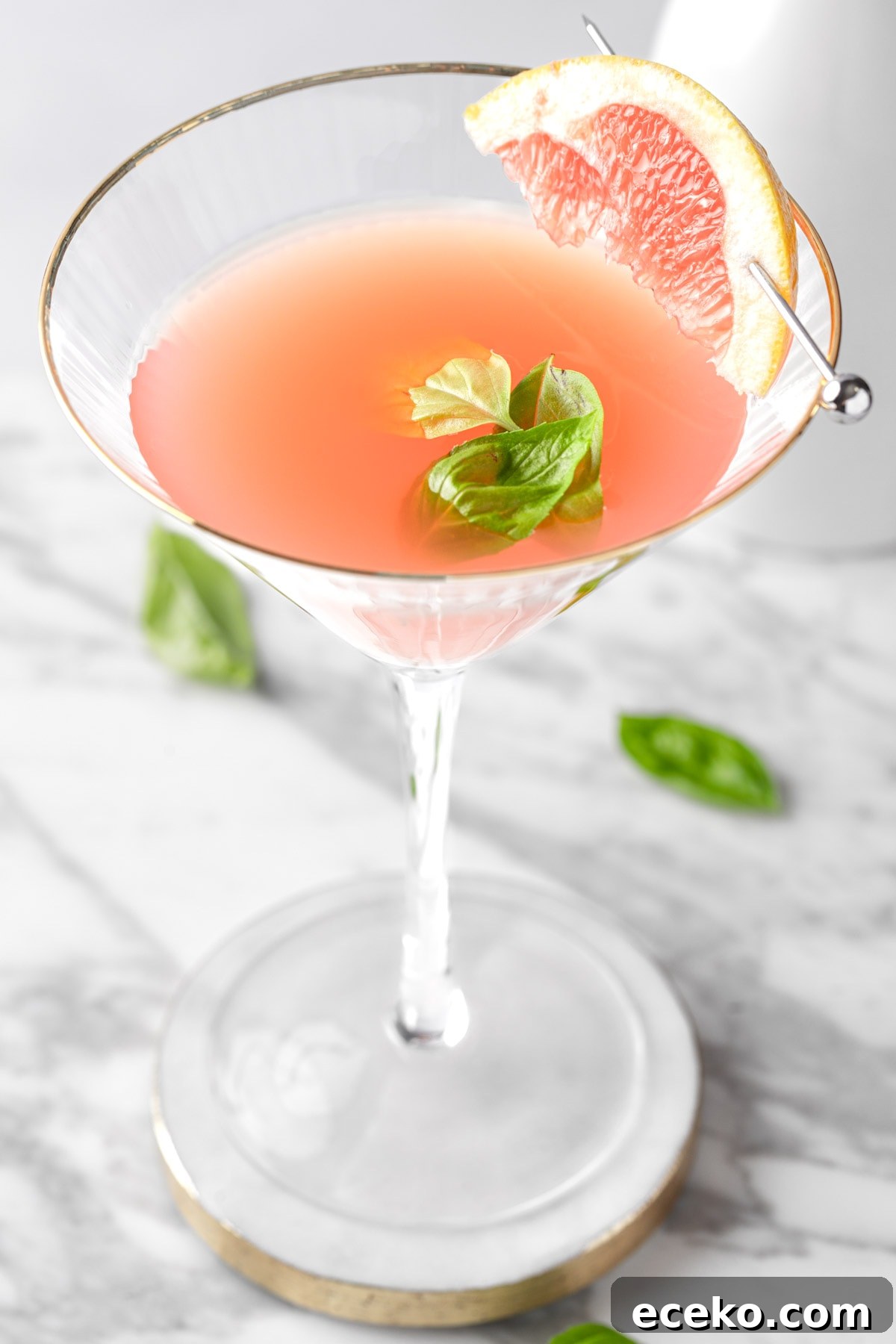 A basil grapefruit martini garnished with a slice of grapefruit, on a white marble table.