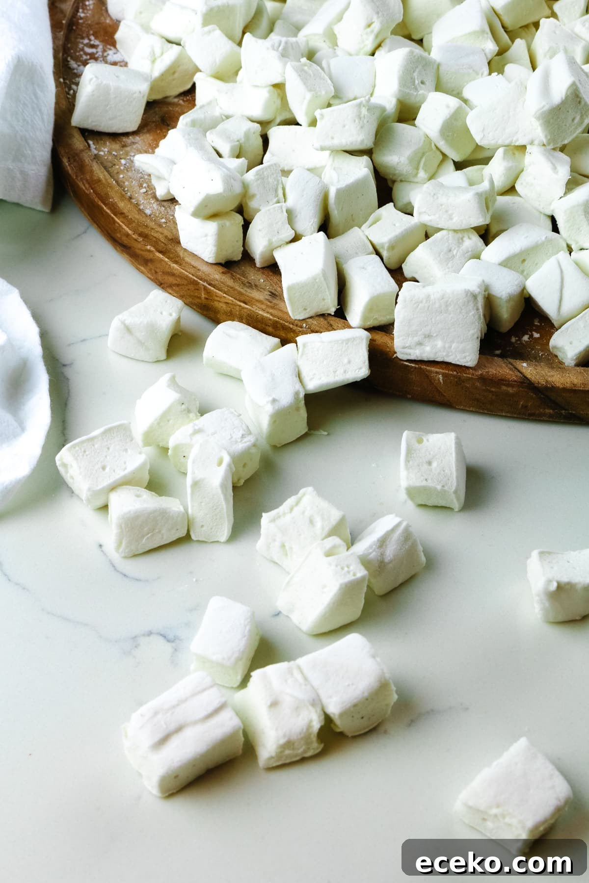 Homemade Marshmallows Without Corn Syrup — Simple Recipe 10 bite-sized homemade marshmallows scattered on a white countertop with a wooden cutting board on the side.