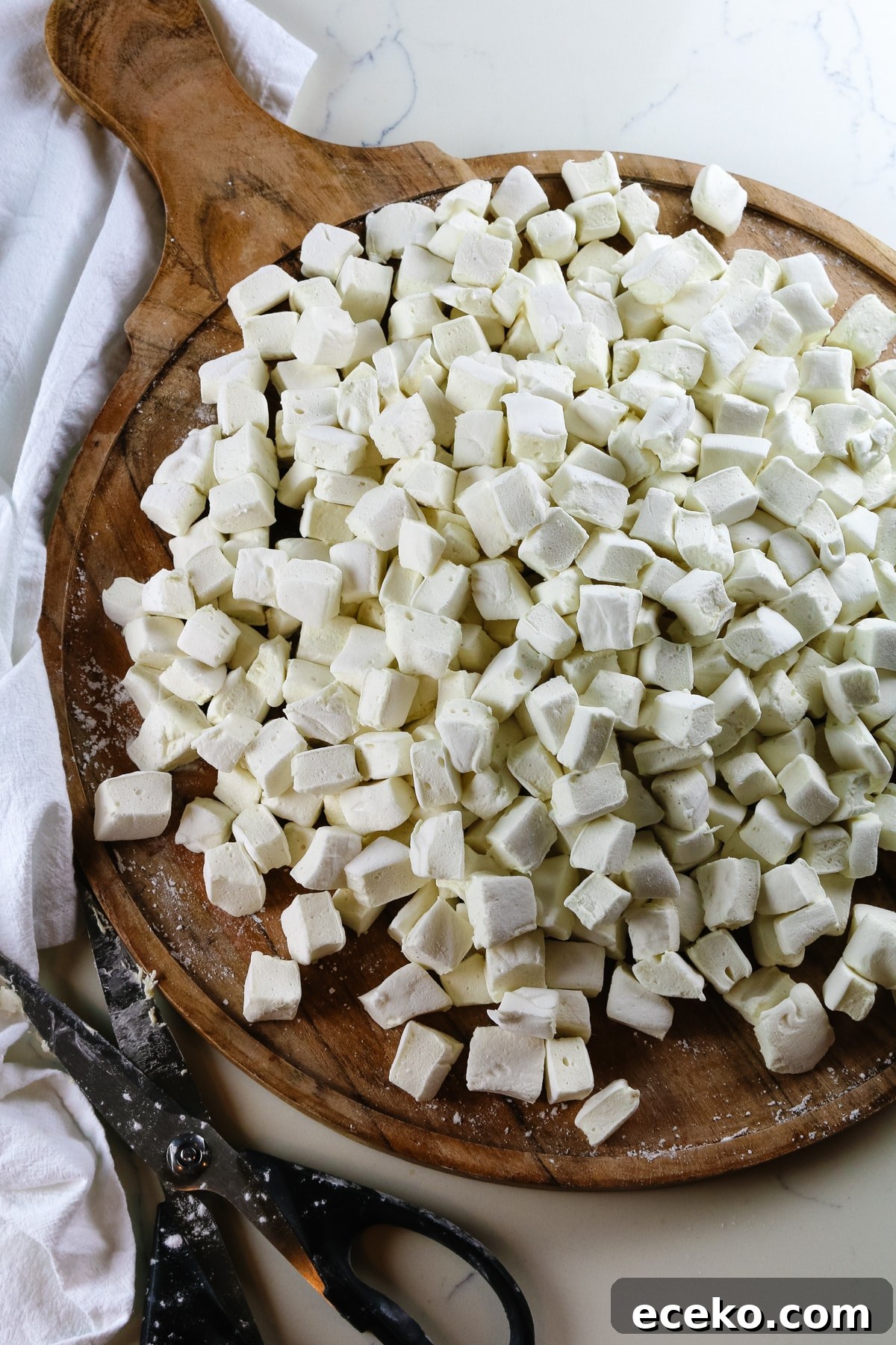 Homemade Marshmallows Without Corn Syrup — Simple Recipe 11 homemade marshmallows cut into bite-size pieces on a circular wooden cutting board and kitchen shears to the side.