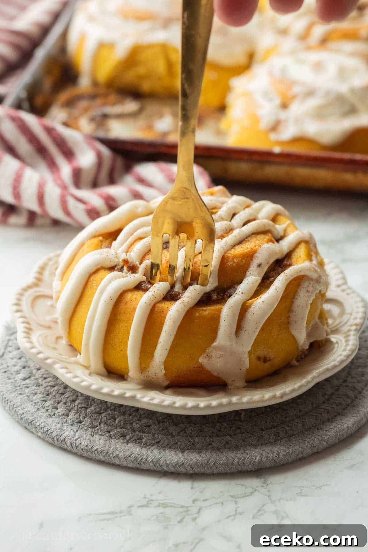 Gold fork in iced pumpkin cinnamon roll on white plate.