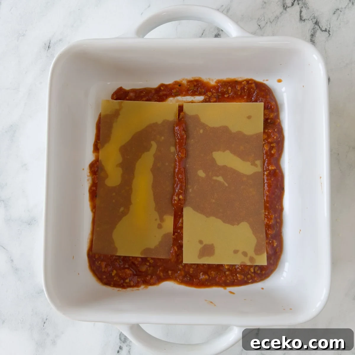 Building lasagna with sauce and noodles in a white square baking dish.