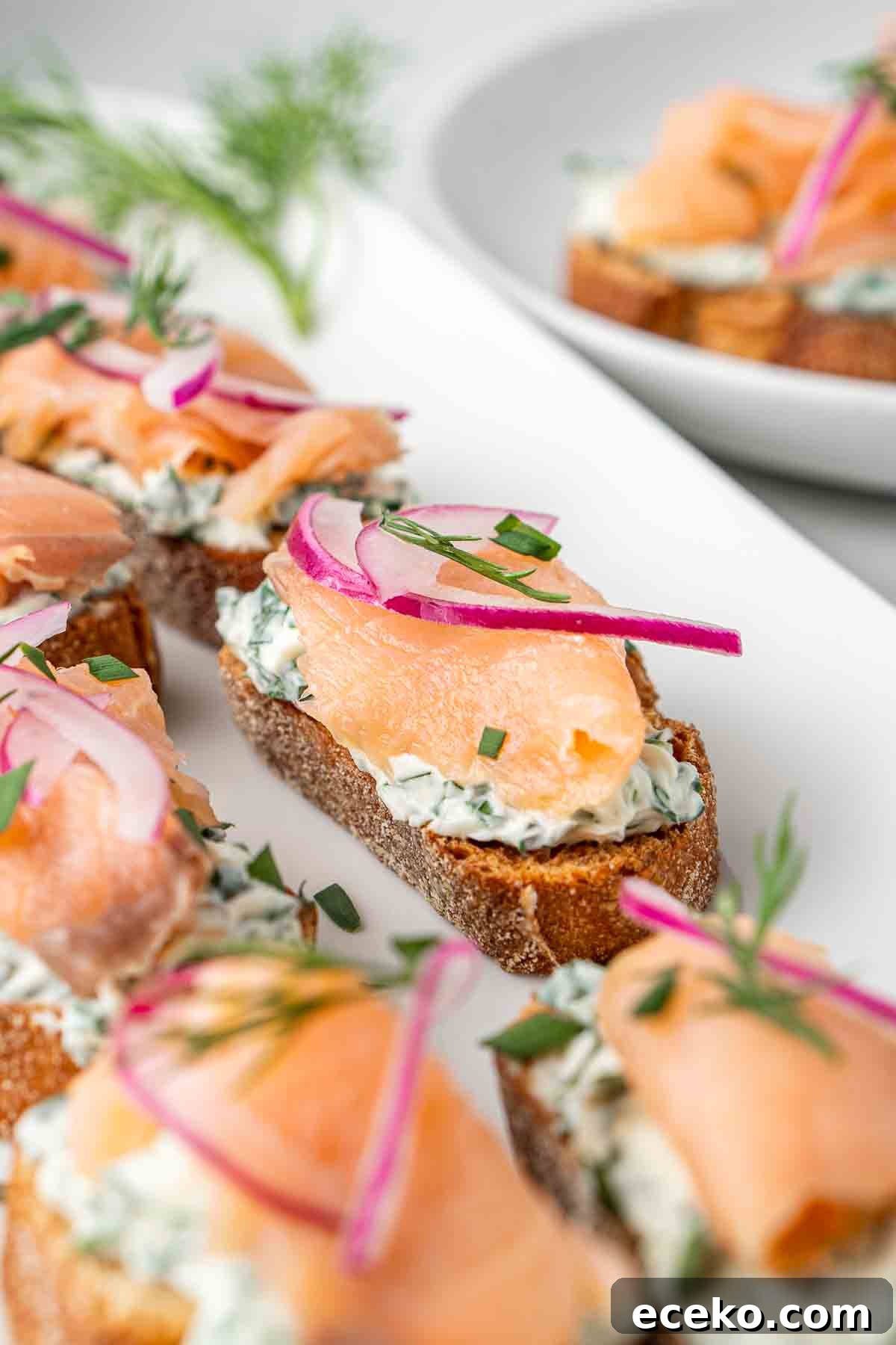 Smoked Salmon Crostini Recipe with Dill Cream Cheese & Capers 8 Close up of smoked salmon crostini on a white serving platter with pickled onion and chives.