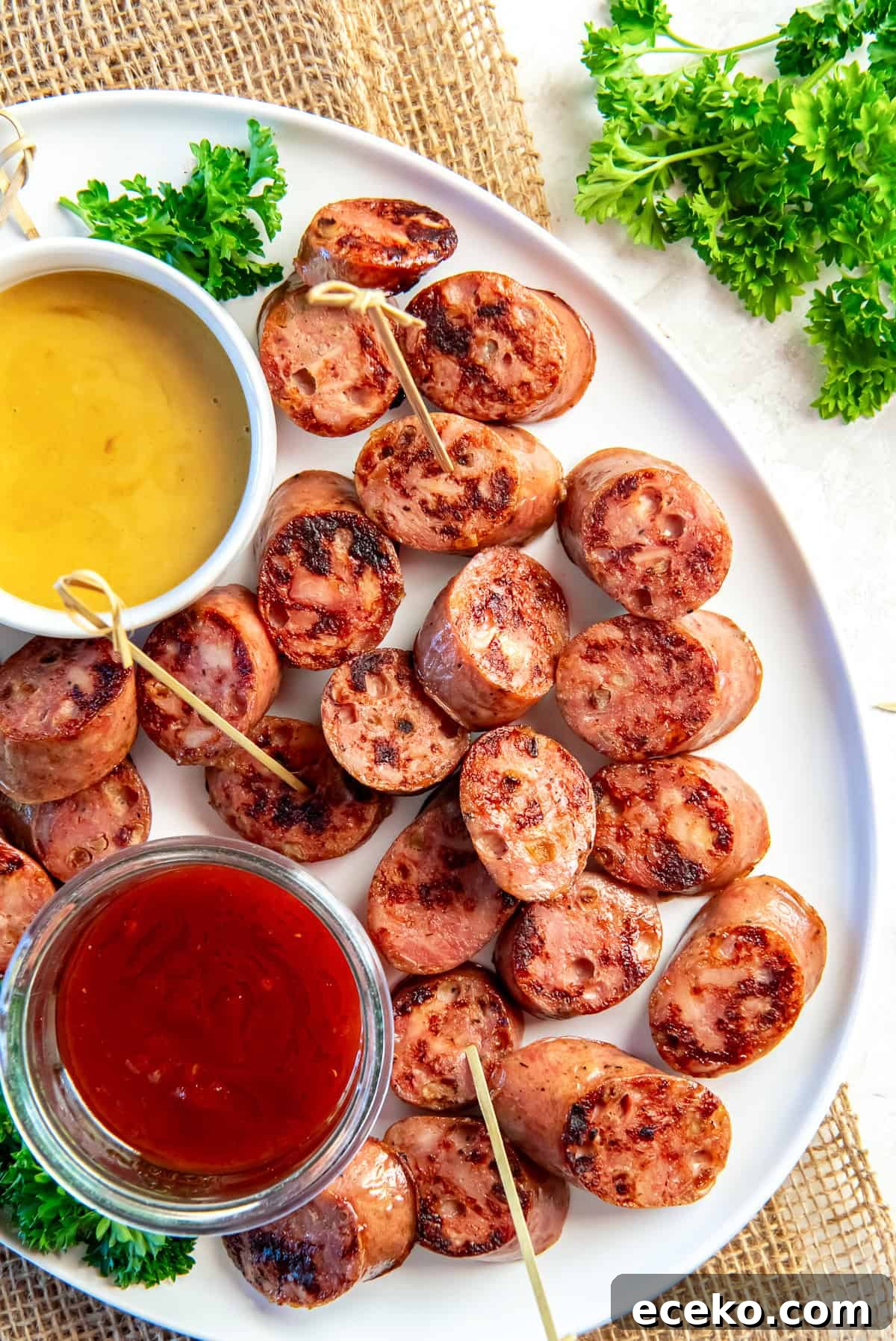 Savory Chicken Sausage Bites for Party Appetizers 2 Chicken sausage bites browned in a skillet and arranged on a white platter with toothpicks, served with honey mustard and chili jam dipping sauces and fresh parsley garnish.