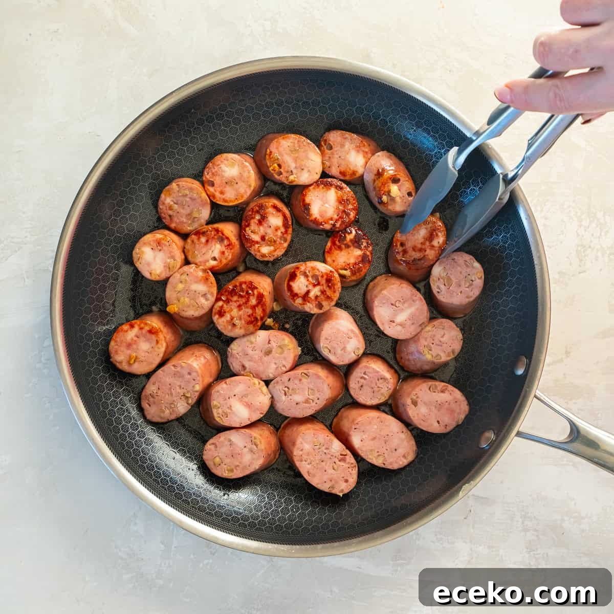 Savory Chicken Sausage Bites for Party Appetizers 6 Overhead view of sliced chicken sausage browning in a large nonstick skillet, with tongs flipping a piece to show golden edges.