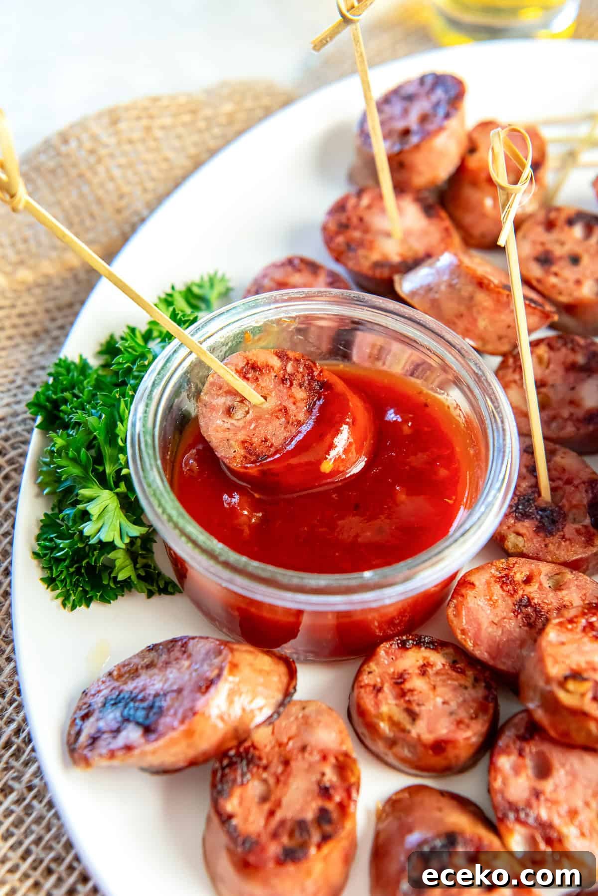 Savory Chicken Sausage Bites for Party Appetizers 7 Chicken sausage bite dipped into a sweet chili jam sauce in a small glass bowl, served on a white platter with toothpicks and fresh parsley.