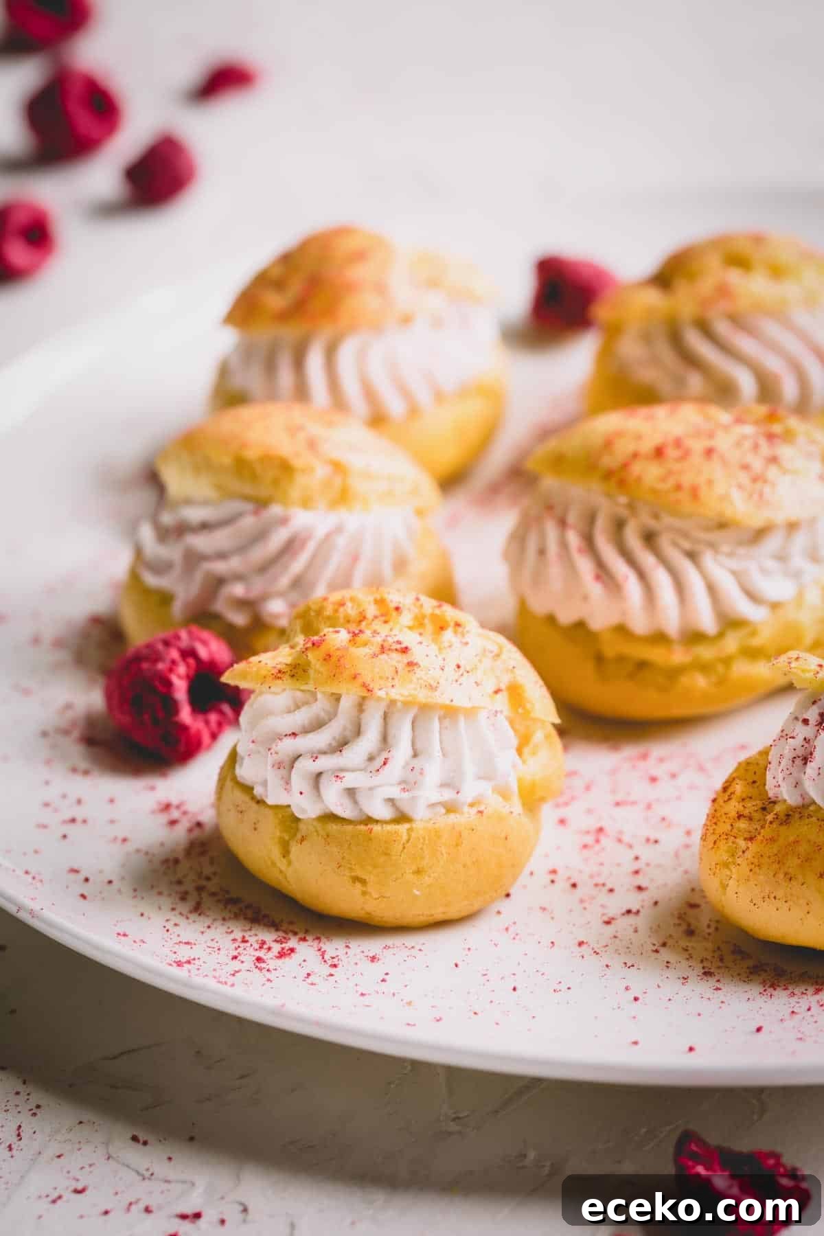 Raspberry Crème Puffs with Vanilla Cream Filling 2 Mini raspberry cream puffs on a white platter dusted with freeze-dried raspberry powder.