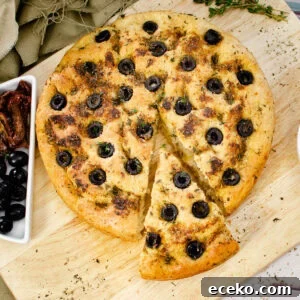 Olive Focaccia Bread on a wooden chopping board with slice cut out served with black olives and sun dried tomatoes on the side