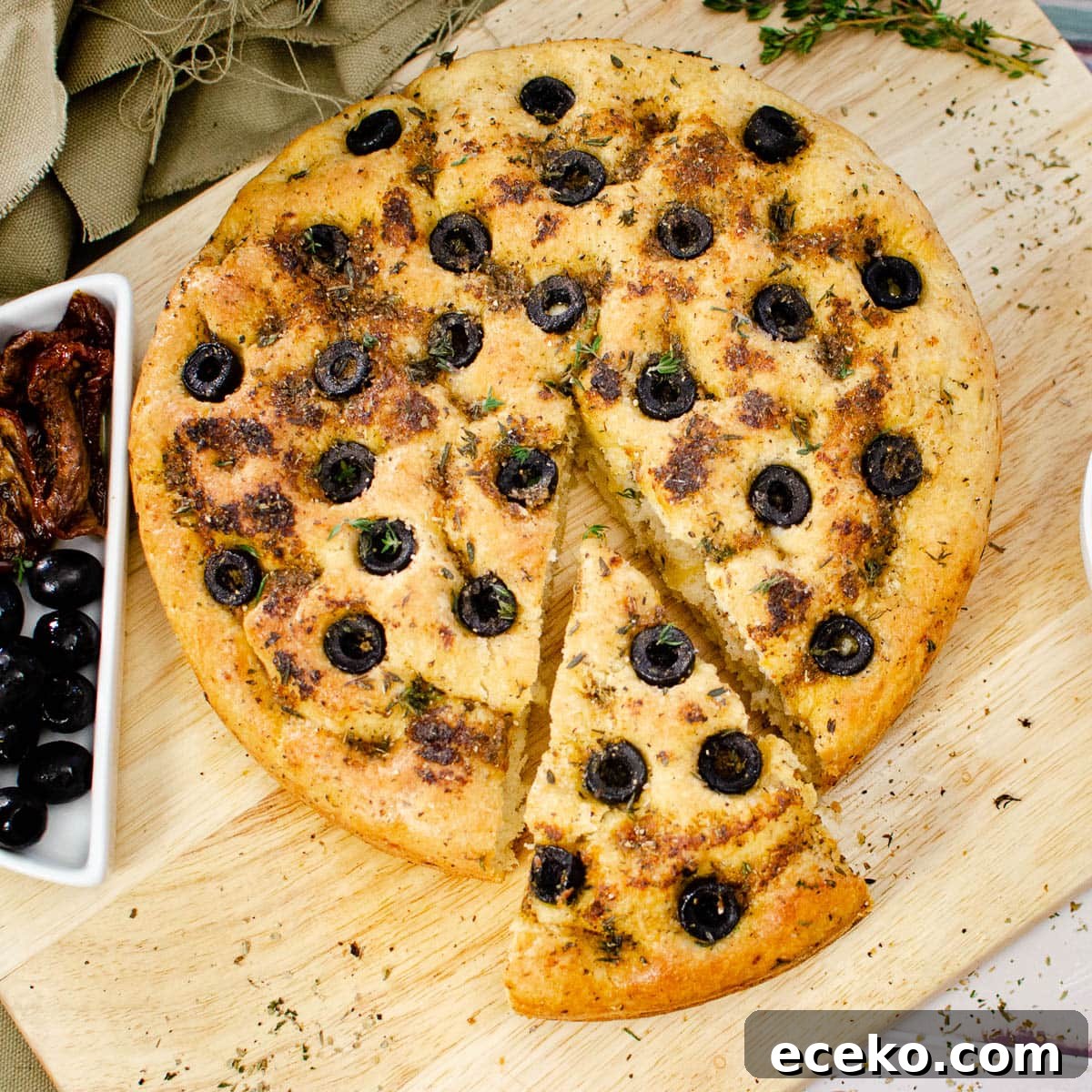 Olive and Herb Focaccia Bread Recipe 5 Olive Focaccia Bread on a wooden chopping board with slice cut out served with black olives and sun dried tomatoes on the side