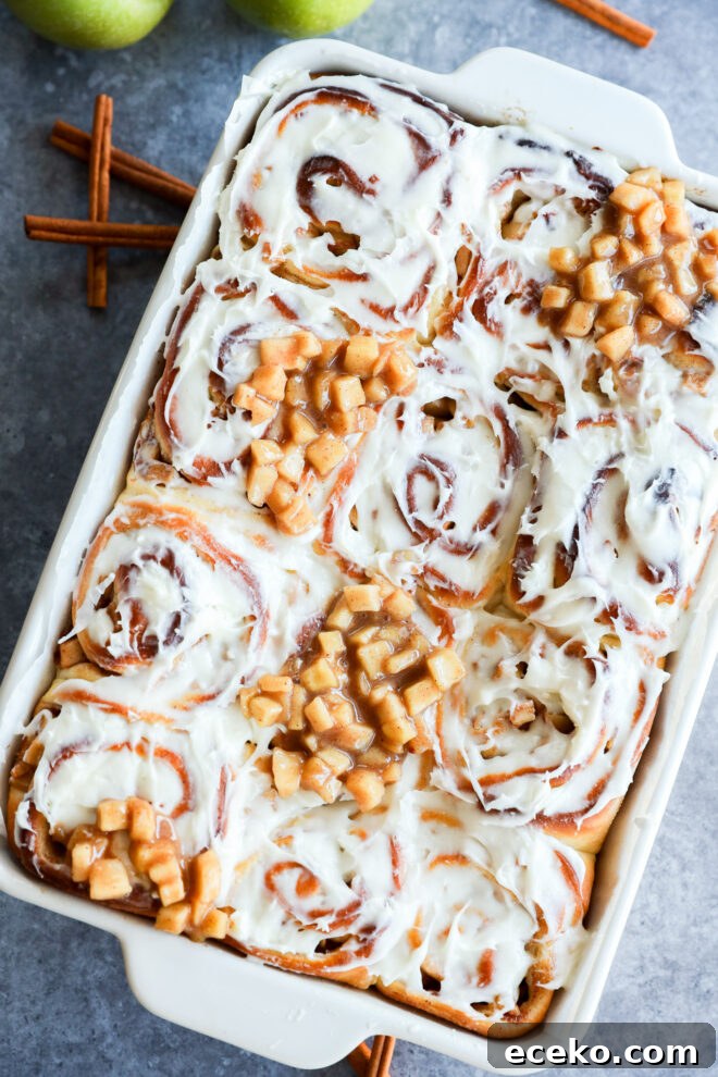 Cinnamon Rolls Stuffed with Apple Pie Filling 2 A baking dish filled with Apple Pie Filled Cinnamon Rolls, some topped with small diced apple pieces. Two green apples and cinnamon sticks are in the background on a gray surface.