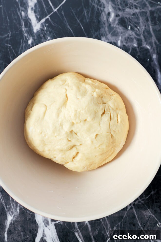 Cinnamon Rolls Stuffed with Apple Pie Filling 3 A ball of dough for Apple Pie Filled Cinnamon Rolls rests in a light-colored mixing bowl, placed on a dark, marbled countertop.