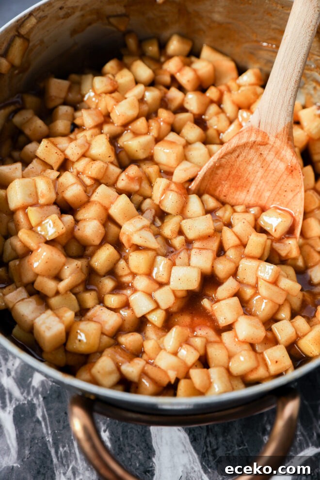 Cinnamon Rolls Stuffed with Apple Pie Filling 4 A saucepan filled with diced apples coated in a glossy cinnamon sauce, reminiscent of Apple Pie Filled Cinnamon Rolls, is being stirred with a wooden spoon on a marble surface.