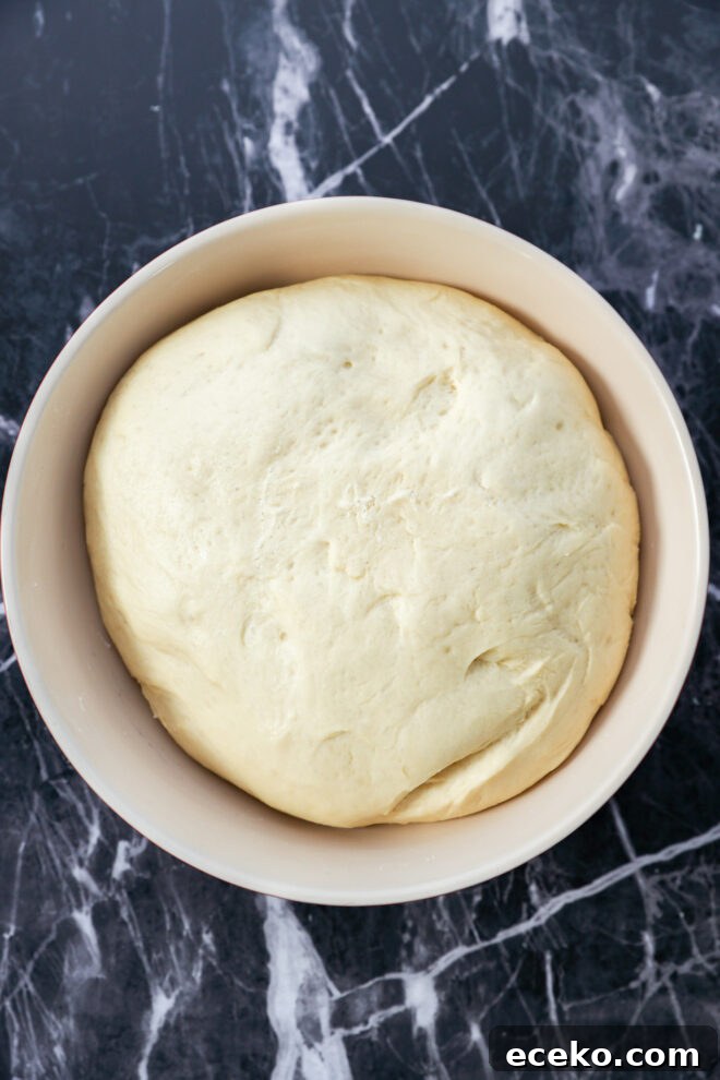 Cinnamon Rolls Stuffed with Apple Pie Filling 5 A large, smooth ball of dough—destined for Apple Pie Filled Cinnamon Rolls—rests in a light-colored bowl on a dark, marbled countertop.