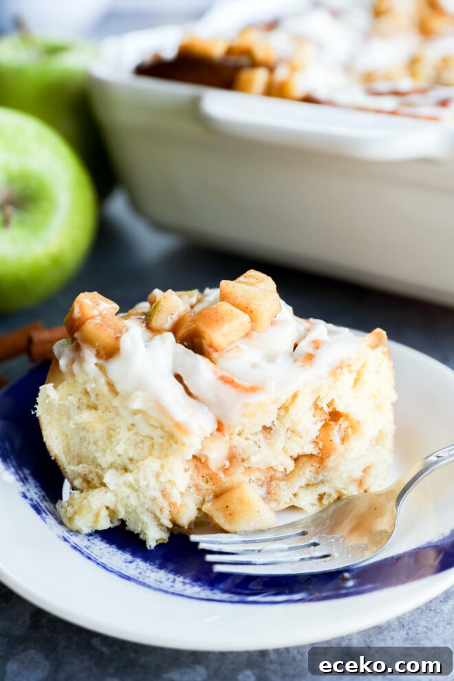 Cinnamon Rolls Stuffed with Apple Pie Filling 9 A close-up of Apple Pie Filled Cinnamon Rolls topped with diced apples and white icing on a blue and white plate with a fork. A baking dish and green apples are visible in the background.