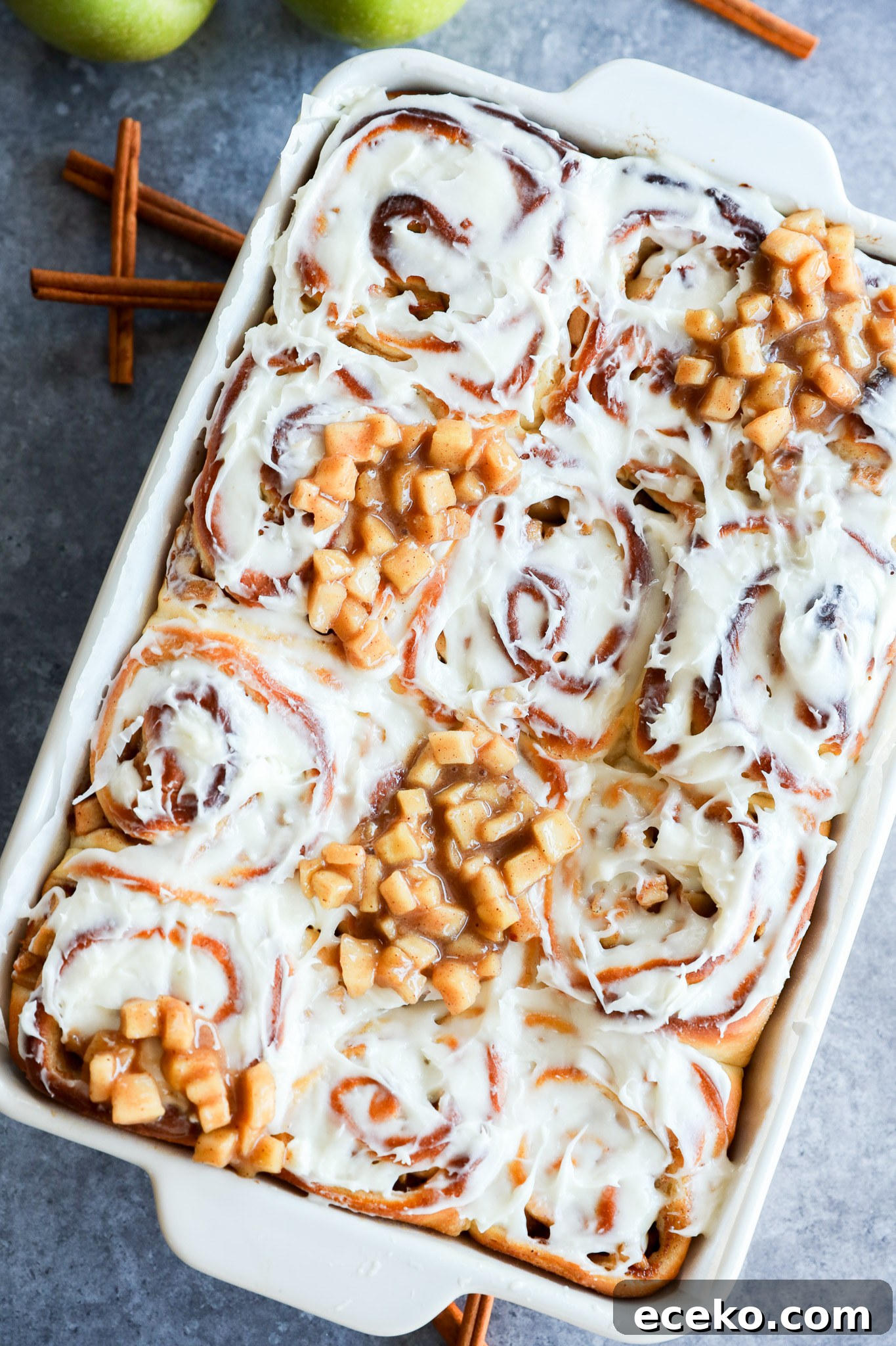 Cinnamon Rolls Stuffed with Apple Pie Filling 10 A baking dish filled with Apple Pie Filled Cinnamon Rolls, some topped with small diced apple pieces. Two green apples and cinnamon sticks are in the background on a gray surface.
